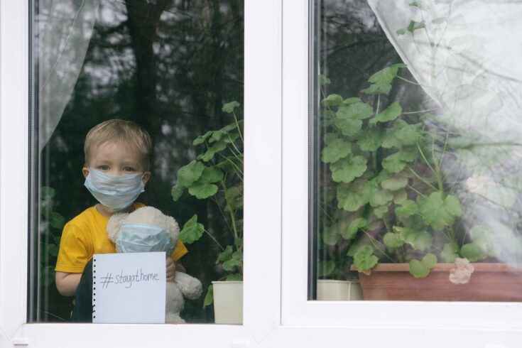 Child in face mask looking out from behind closed window