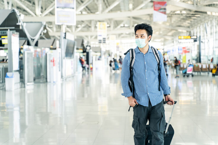 Asian traveler with luggage, wearing a face mask and looking at the time table in the departure terminal in an airport during the COVID-19 virus pandemic.