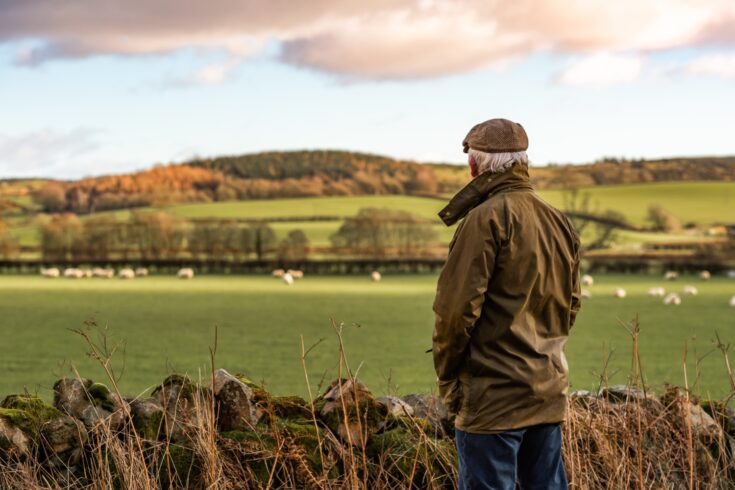 A man with his back to camera wearing a waxed jacket and flat tweed cap looking across a field