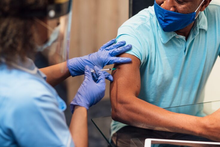 A mixed race female nurse wearing a protective face shield, surgical mask and protective gloves administering the COVID-19 vaccine to a black man