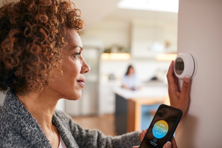 Woman using app on smart phone to control digital central heating thermostat at home