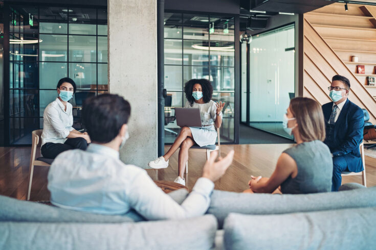 Group of entrepreneurs, wearing masks, sitting at a distance in the office and talking.