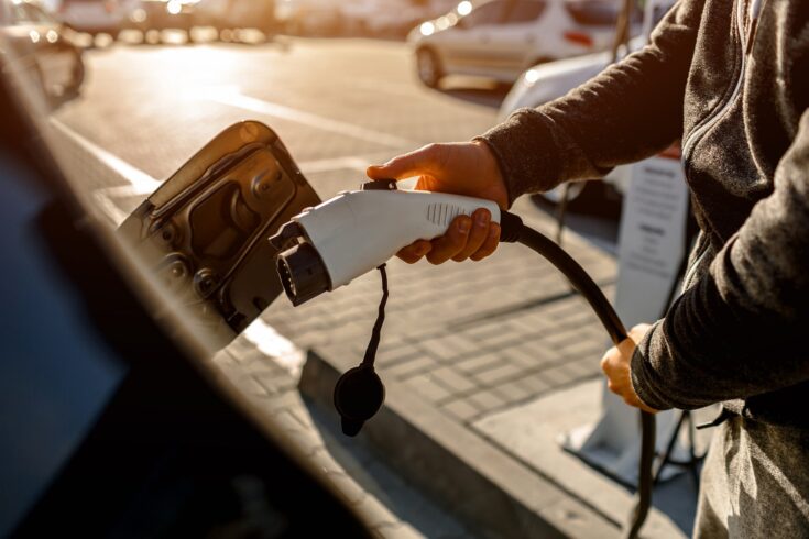 Man Holding Power Charging Cable For Electric Car In Outdoor Car Park. And heâs going to connect the car to the charging station in the parking lot near the shopping center