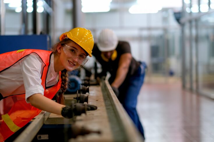 Female technician smile and look forward in front of rail of the machine with her co-worker as background in factory