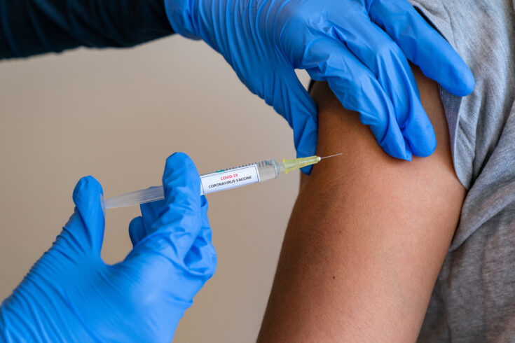 Female doctor wearing blue latex gloves injecting a child in her arm with a needle and syringe containing a dose of the COVID-19 vaccine cure by way of immunisation