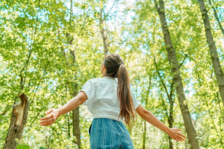 Free woman breathing clean air in nature forest. Happy girl from the back with open arms in happiness. Fresh outdoor woods, wellness healthy lifestyle concept.