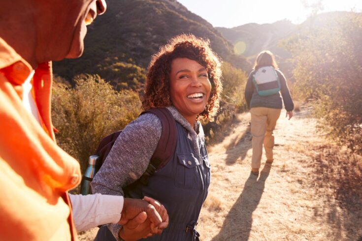 Woman Helping Man On Trail As Group Of Senior Friends Go Hiking In Countryside Together