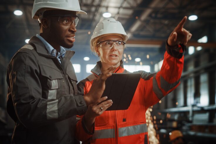 Two Heavy Industry Engineers Stand in Steel Metal Manufacturing Factory, Use Digital Tablet Computer and Have a Discussion. Black African American Industrial Specialist Talk to Female Technician.