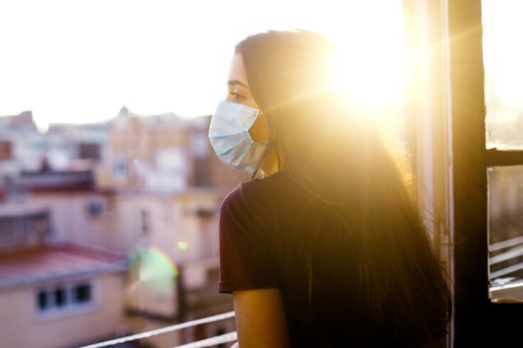 Teenage girl in quarantine wearing protective mask looking out the window at sunset