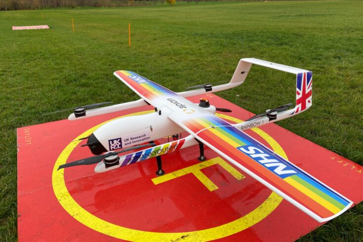 A fixed wing drone with rainbow coloured wings sits on a red square helipad in a field