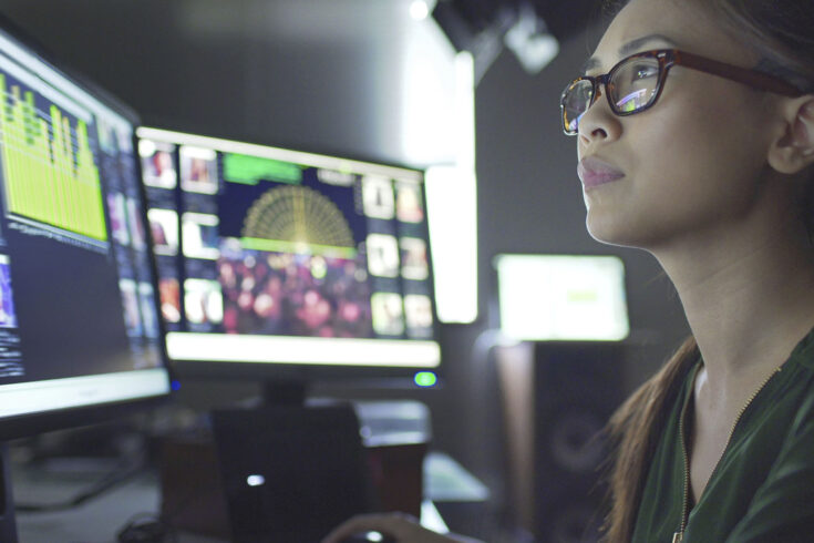 Close up of a young Asian woman sitting down at her desk where she’s surrounded by 3 large computer monitors displaying out of focus images of people as thumbnails, crowds, graphs and scrolling text.
