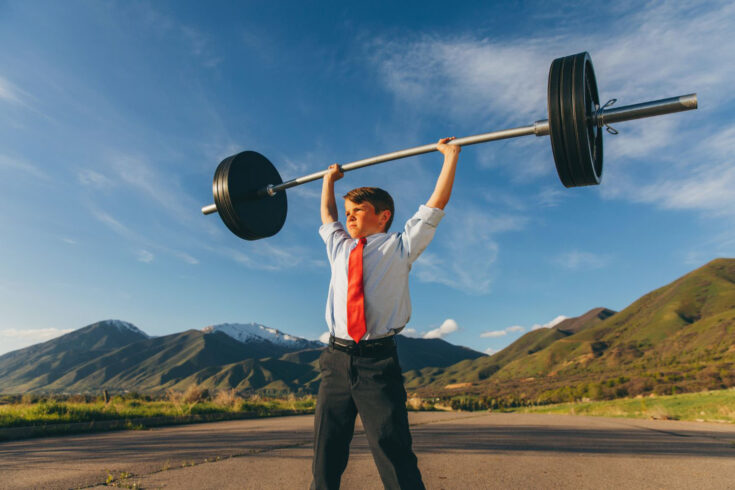 A young boy dressed as a businessman is showing his strength outside by lifting a barbell and weights. He is showing his confidence and inner strength to accomplish a difficult task amidst adversity. Image taken in Utah, USA.