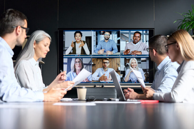 Online videoconference in meeting room with diverse people sitting in modern office and multicultural colleagues on big screen monitor.