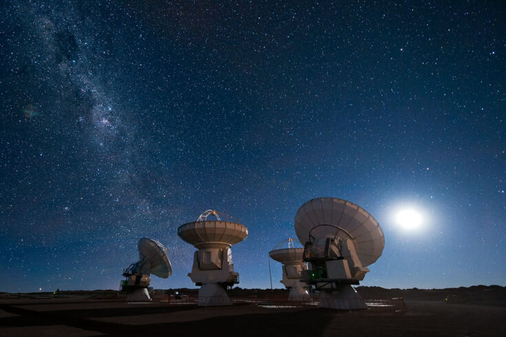 Four antennas of the Atacama Large Millimeter or submillimeter Array (ALMA) gaze up at the star-filled night sky, in anticipation of the work that lies ahead. The moon lights the scene on the right, while the band of the Milky Way stretches across the upper left.