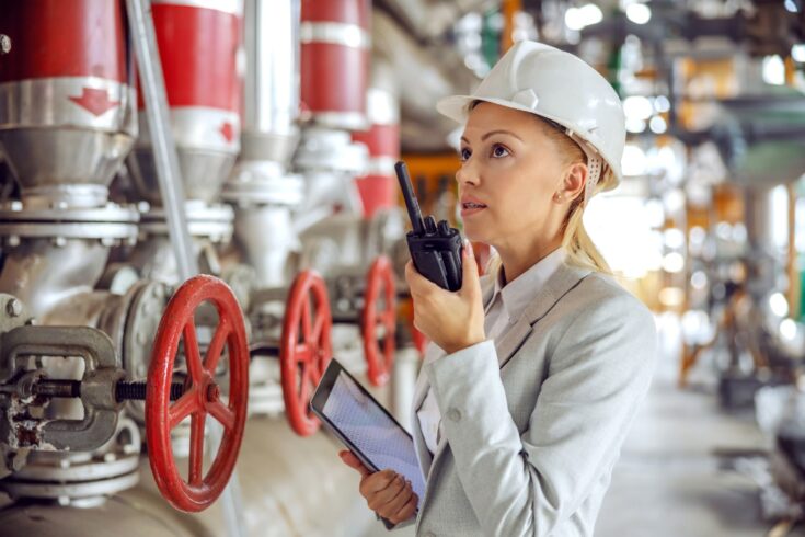 A female supervisor with helmet in suit holding tablet in hands and talking on walkie talkie while standing in a heating plant