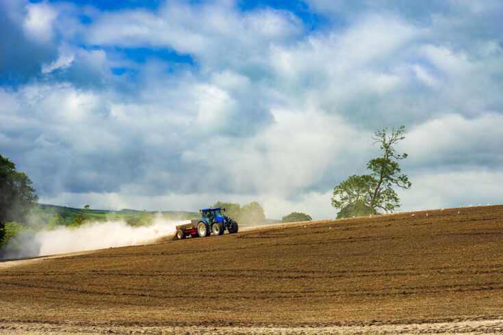 A cloud of dust expands behind a tractor ploughing a field.