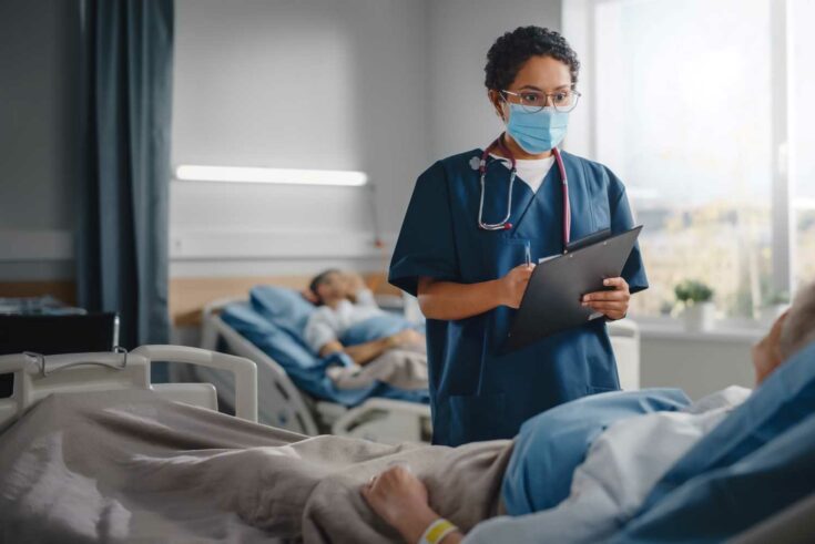 Hospital Ward: Friendly Nurse Wearing Face Mask Talks to Patient Resting in Bed