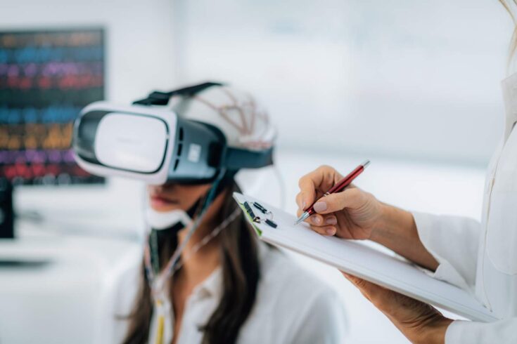 A patient in a neuroscience lab with virtual reality goggles and EEG brainwave cap. Another person is taking notes on the side.