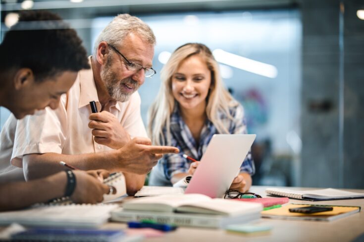 Happy mature professor and his students using laptop at campus