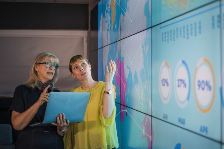 An experienced woman mentors a female colleague, the mature woman is holding a laptop as they debate data from an interactive display; they are both wearing smart casual clothing.