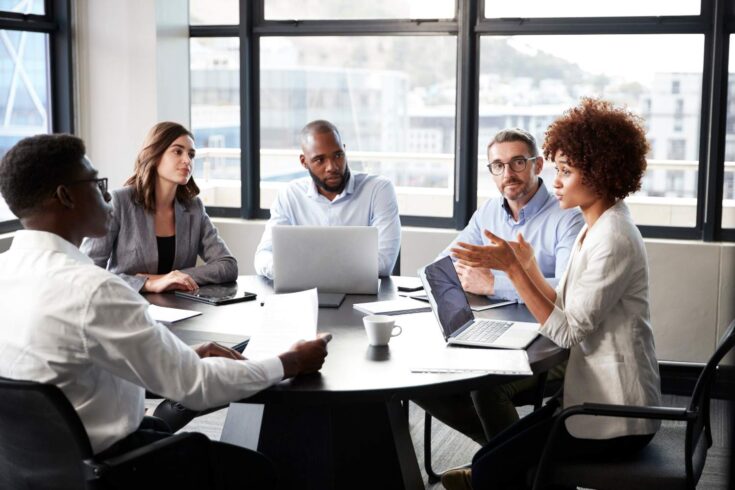 Diverse group of people sitting around table with their laptops discussing at workplace