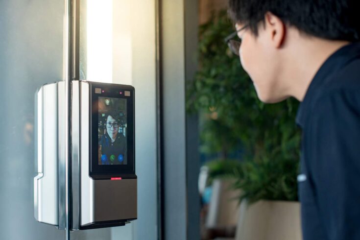 Asian man using face scanner to unlock glass door in office building.