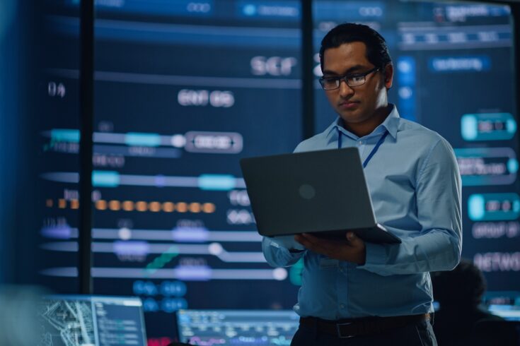 Young multiethnic male employee uses laptop computer in system control monitoring centre