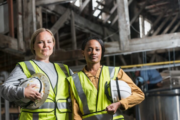 Two multi-ethnic women working in a warehouse holding white hardhats and wearing yellow safety vests. They are looking at the camera.