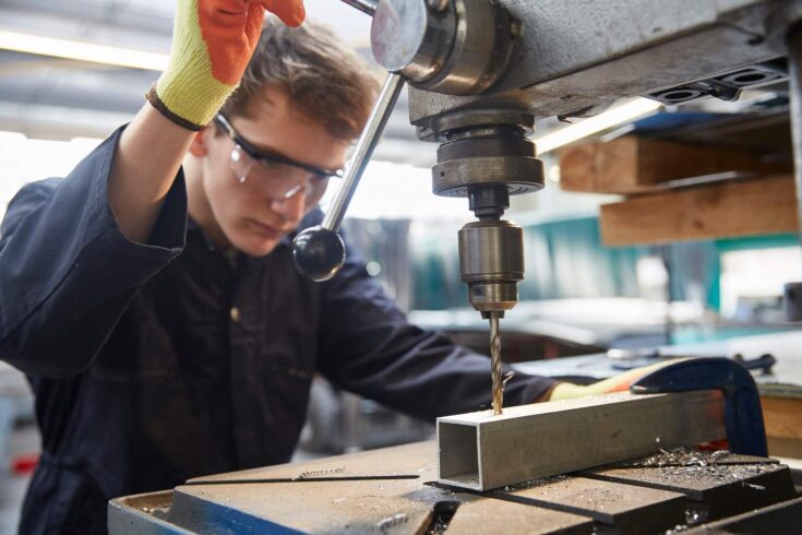 Young apprentice in safety goggles using pillar drill in steel fabrication factory