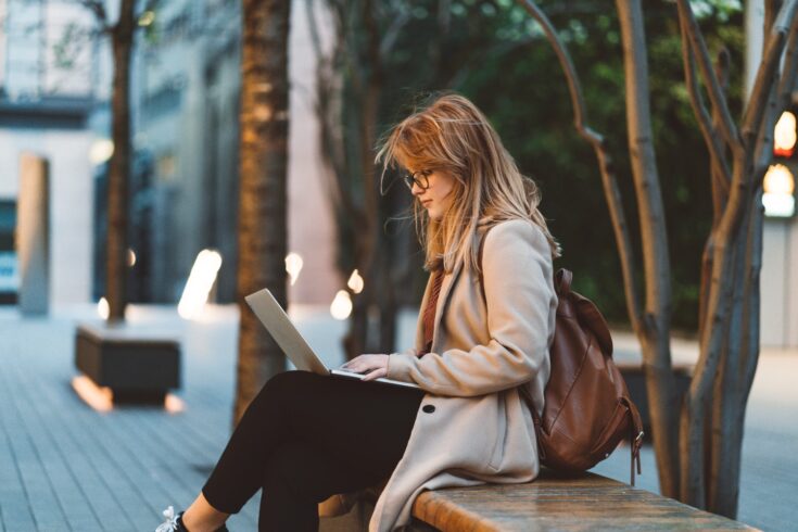 Young businesswoman outside, walking, having a phone conversation, working, having a video call. Student sitting on a bench in a park while using laptop and mobile phone at dusk.