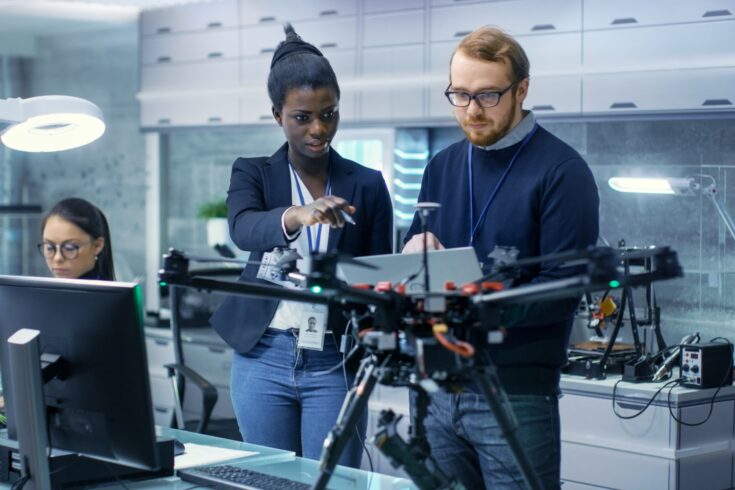 Caucasian Male and Black Female Engineers Working on a Drone Project with Help of Laptop and Taking Notes.