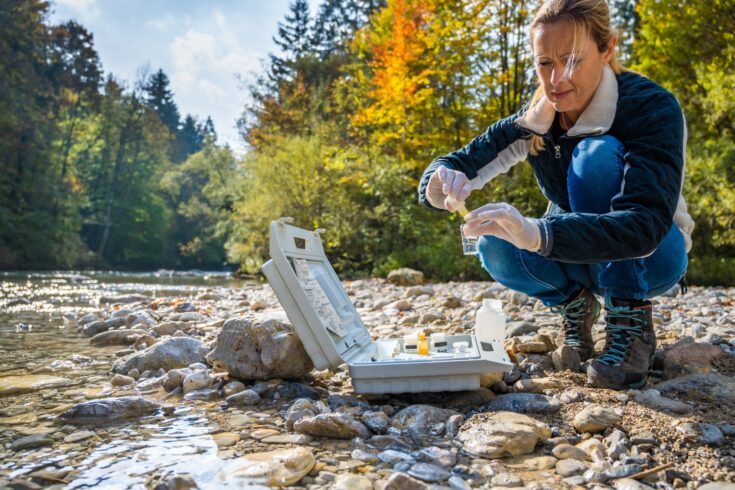 Mature female biologist adding reagent into a vial with water sample near river.