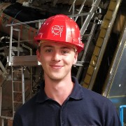Alex Headspith wearing a red CERN hat. Machinery from CERN in the background.