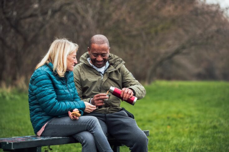 A senior mixed race couple wearing warm clothing and sitting on a park bench on a day in December. They are having a snack and a warm drink out of a flask.