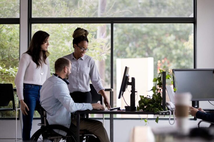 A small group of diverse coworkers using a desktop computer in a large, open-space office.