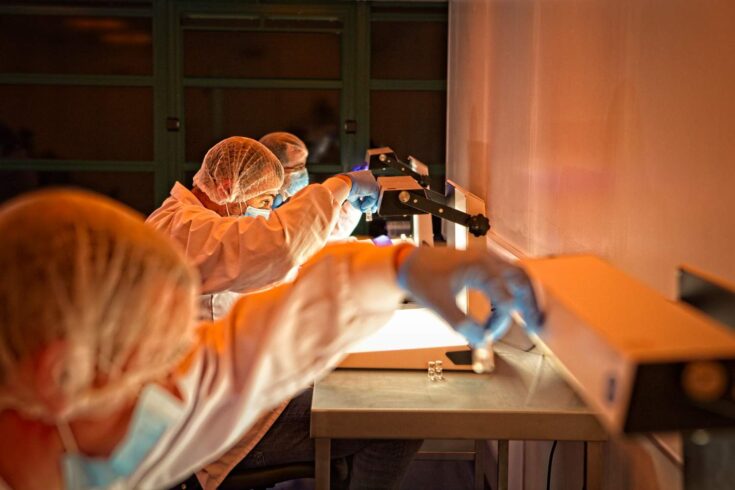 People wearing masks, hair nets and surgical gloves looking at tiny glass containers for medical research purposes.