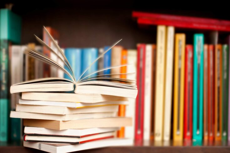 Stack of books on a wooden library shelf, one of them open on top, multicolored book spines background.