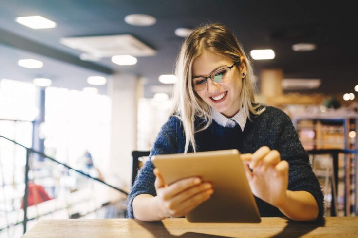 A young woman with glasses smiling and reading an electronic tablet