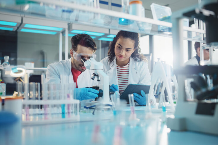 Modern medical research laboratory: portrait of latin and black young scientists using microscope, digital tablet, doing sample analysis, talking. Diverse team of specialists work in advanced lab.