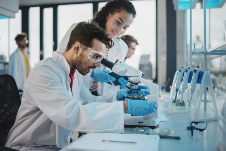 Scientists Using Microscope, Digital Tablet, Doing Sample Analysis, Talking. Diverse Team of Specialists work in Advanced Lab