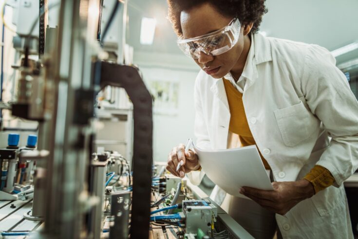 Black female quality control inspector examining machine part on production line.