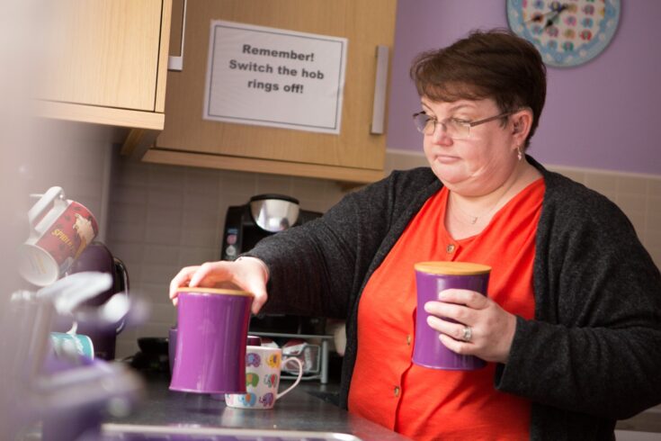 Woman making a cup of tea, sign in background says Remember! Switch the hob rings off.