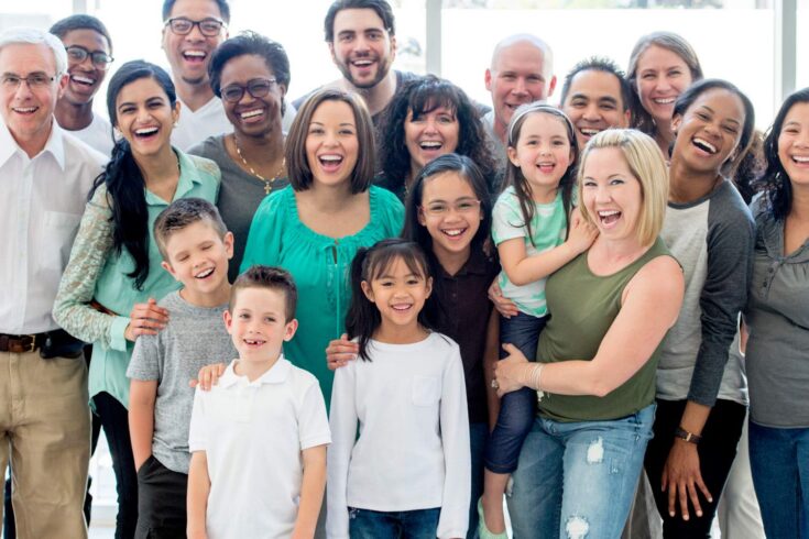 A multi-ethnic family group standing together happily while smiling and looking at the camera.