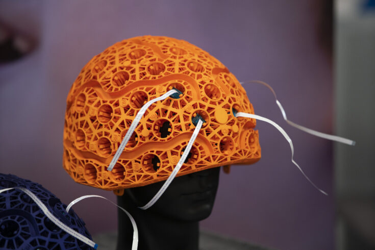 An orange helmet with holes for sensors sits atop a model head. White tape comes through four of the holes.