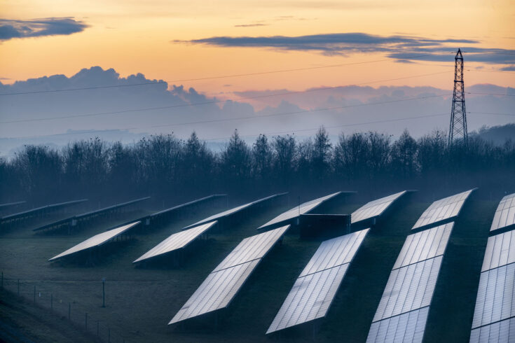 Multiple solar panels reflecting the fading winter sunlight at dusk, and glowing afterlight in the sky beyond, in the rural countryside of southern Britain.