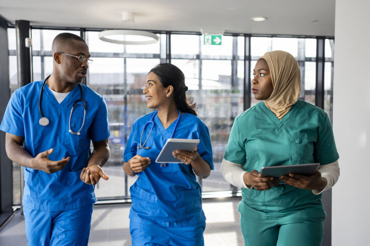 Mixed ethnic group of medical professionals walking down a corridor together in the North East of England. They are working a shift at a hospital and are dressed in scrubs. The women are carrying digital tablets.