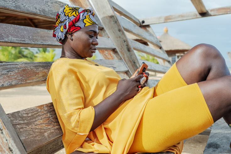 Ghana woman with african colorful headdress sitting on a bench in the village of keta with cellphone.