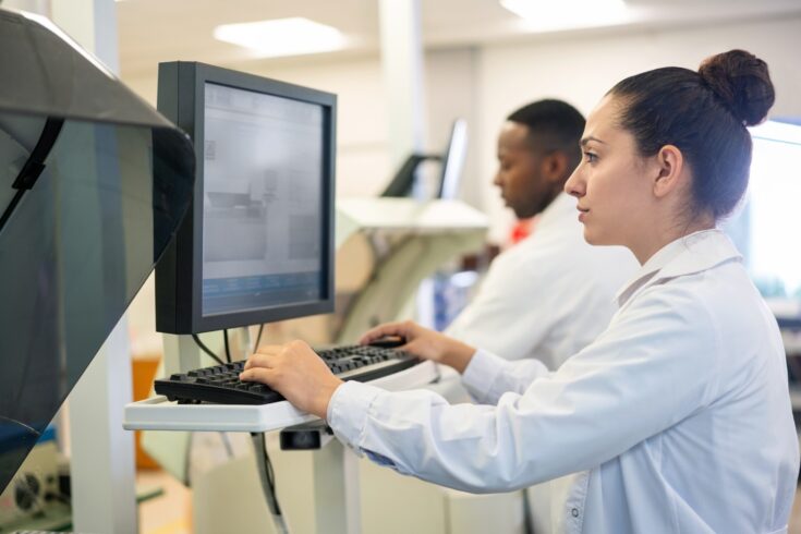 Side view of a woman scientist working on computer with male colleague in background. Medical researchers wearing lab coats working in a modern laboratory.