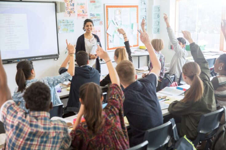 Rear view of teenage students raising hands in classroom