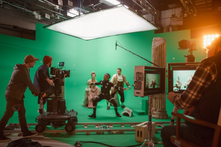 People on set in front of a green screen with a actor in period clothing protecting an actress from a man wearing a motion capture suit